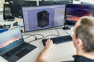 A man sits at his desk at a computer and develops a battery system using CAD software.