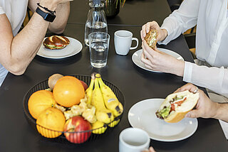 Three people sit at a table set with a fruit basket and crockery and eat sandwiches