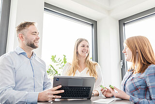 Two women and a man holding a laptop sit at a table during a team meeting